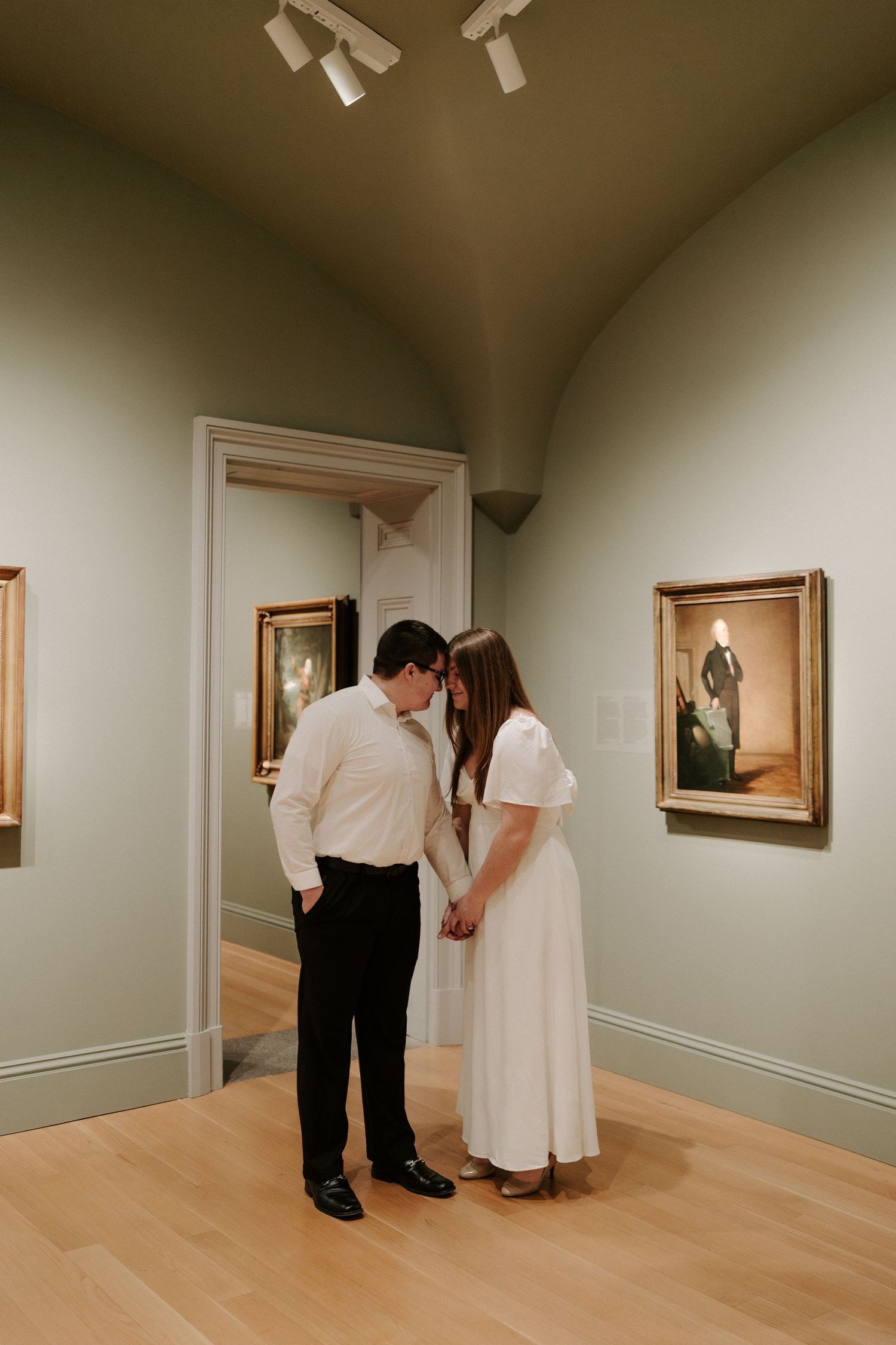 Couple holds hands and shares a quiet moment in a sage green museum gallery surrounded by portraits.