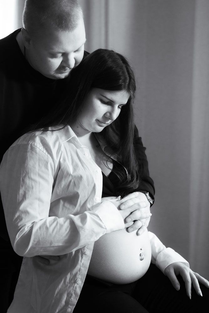 Raskaana oleva nainen istuu tuolilla ja hänen mies seisoo hänen selän takana. He pitävät yhdessä mahasta kiinni.