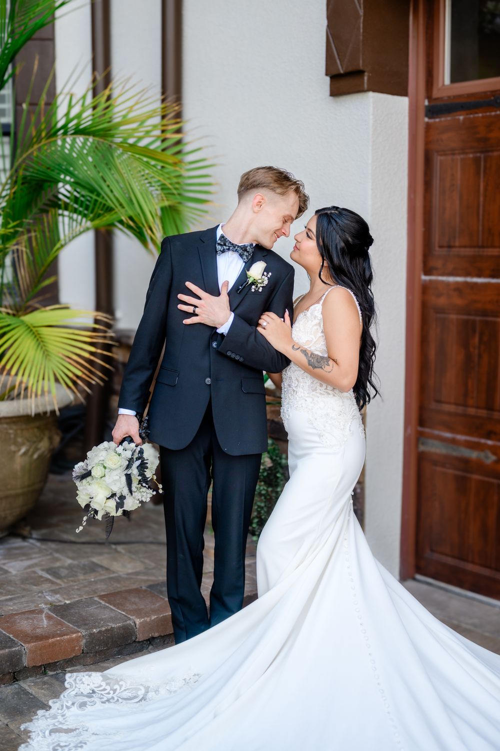 A romantic wedding couple embraces in front of wooden doors and palm trees, with the bride's white dress flowing.