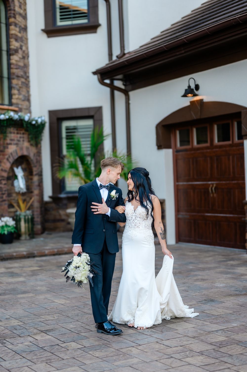 A couple in wedding attire shares a romantic moment on a stone paved driveway with white buildings in the background.
