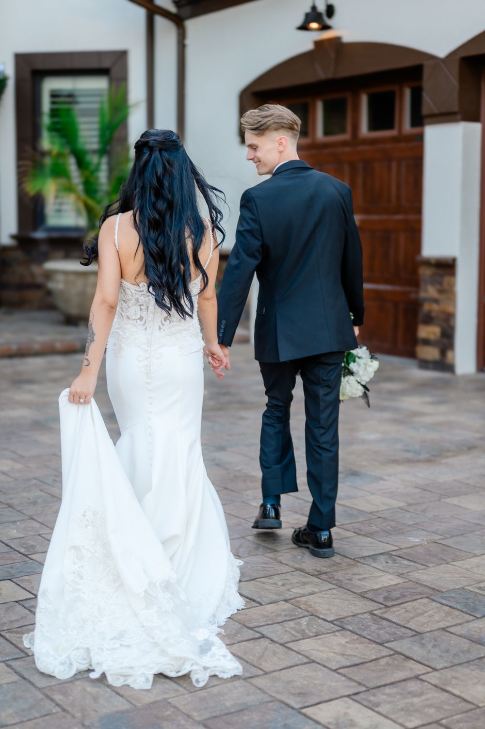 A bride and groom walk together on a stone patio, with her flowing white dress trailing behind them.