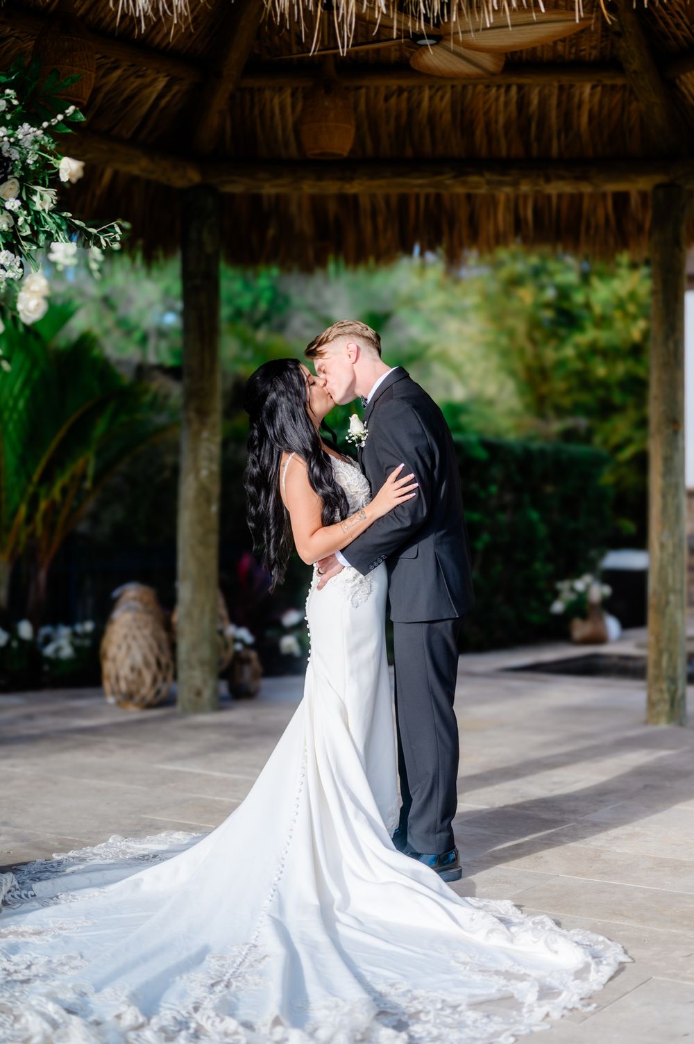 A romantic wedding couple shares a tender embrace under a thatched roof pavilion with flowing white dress details.