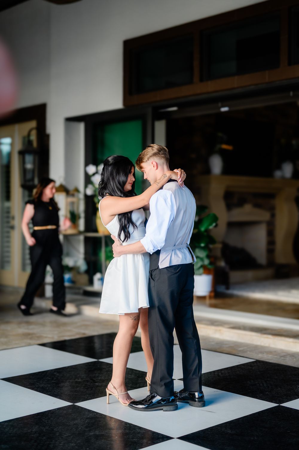 A couple dances together on a black and white checkered floor in an elegant indoor space.