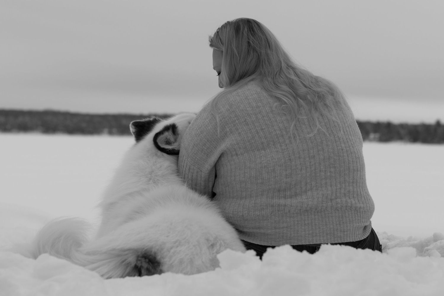 Black and white photo of a woman sitting in snow, embracing a large white fluffy dog outdoors.