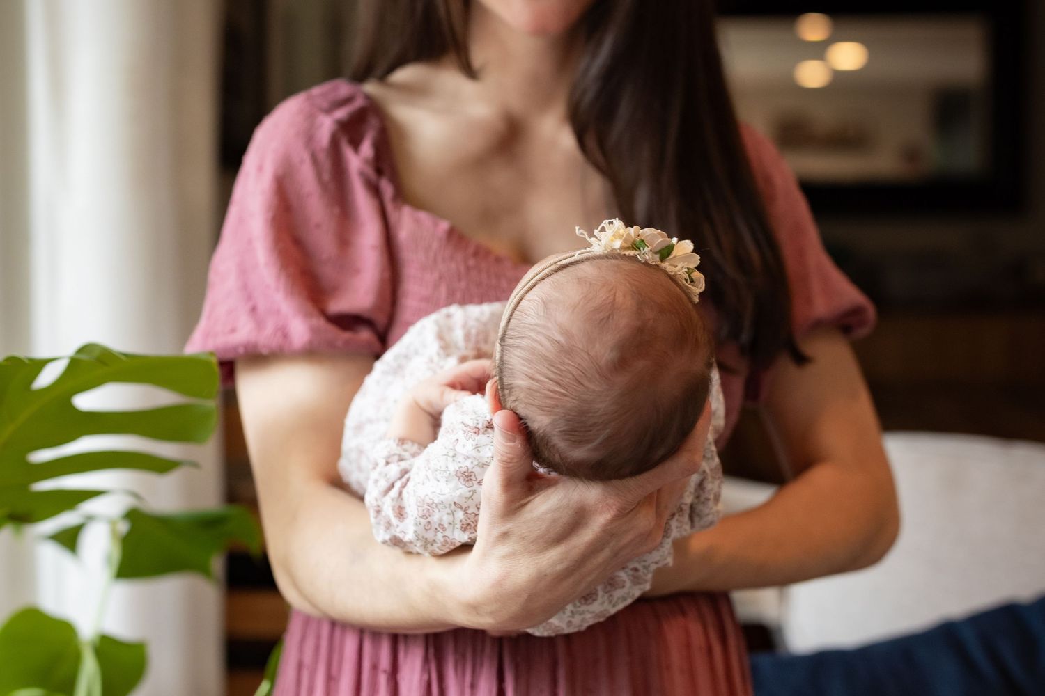 Mother in pink dress tenderly holds newborn baby with floral headband against her chest.