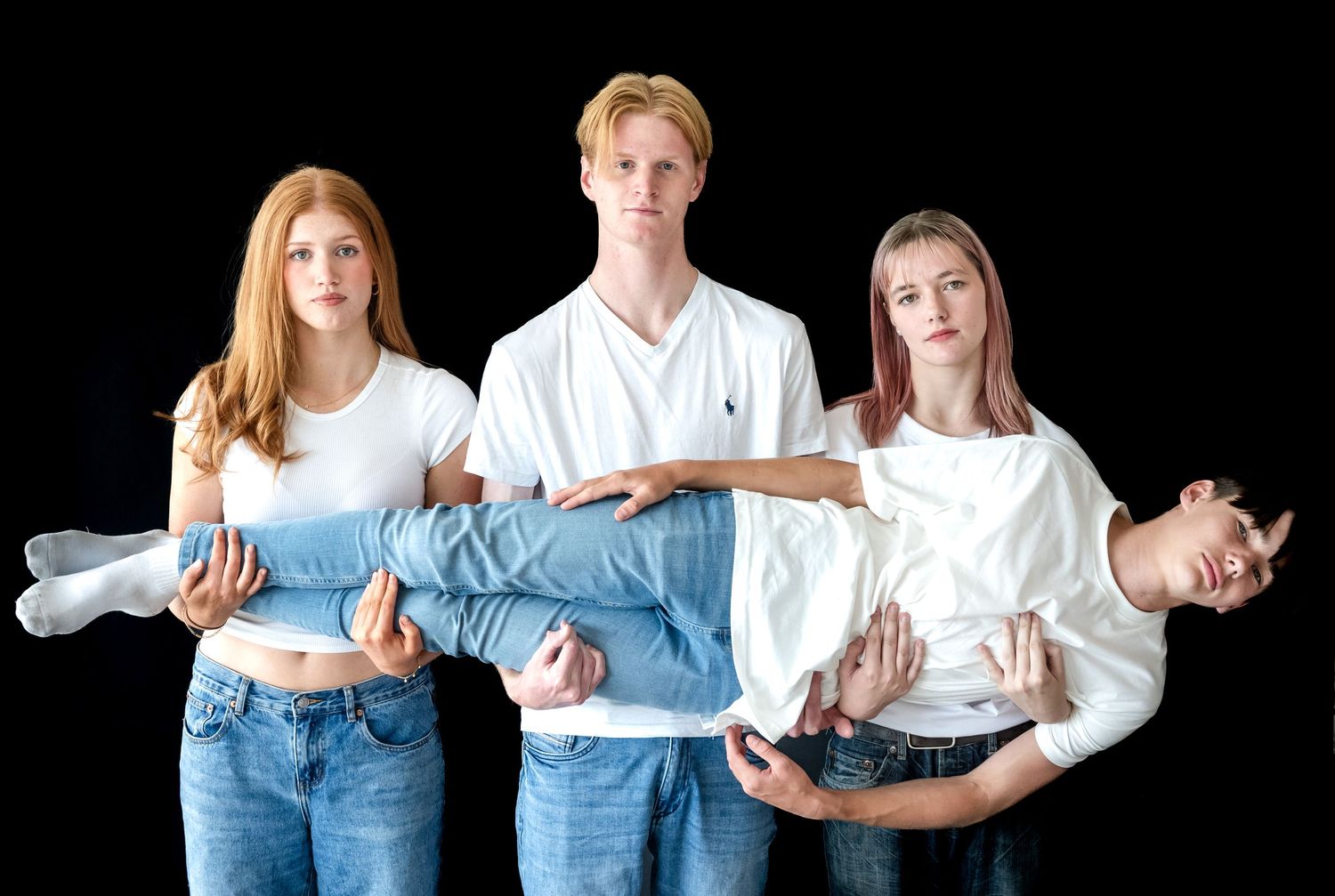 Three people in white shirts and jeans stand together holding someone horizontally in a playful group pose against black background.