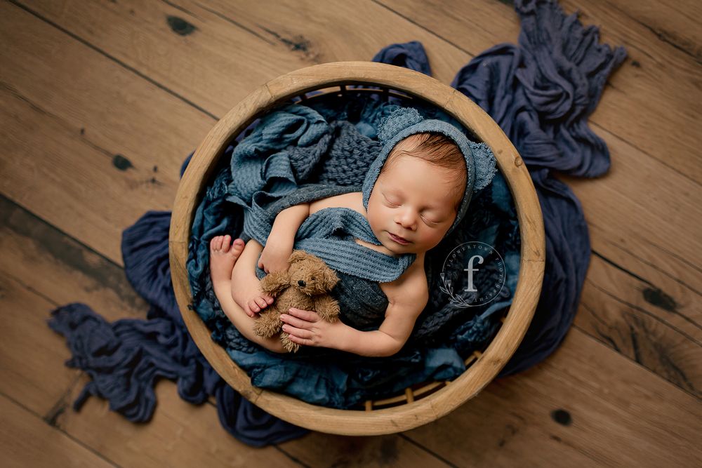 Newborn baby boy sleeping in wooden bowl with blue wrap and hat and teddy bear on rustic wooden floor at Summerville SC newborn studio