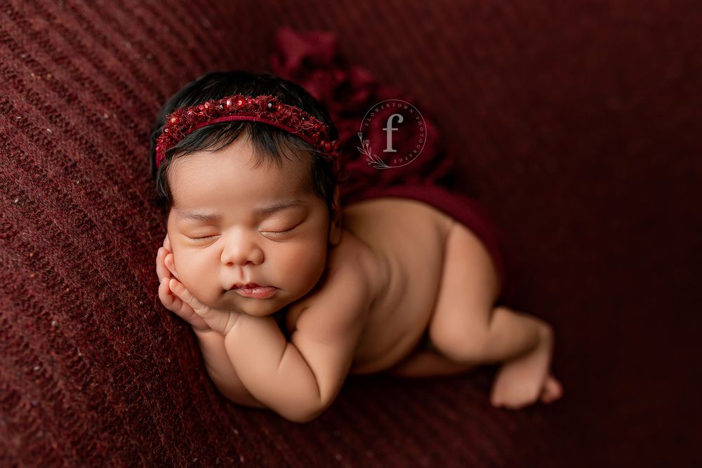 Newborn baby girl with burgundy headband sleeping on rich red knit blanket during studio portrait