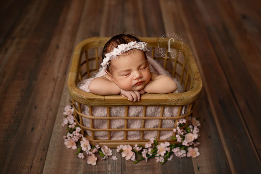Newborn baby sleeping in bamboo basket surrounded by pink blossoms on wood floor