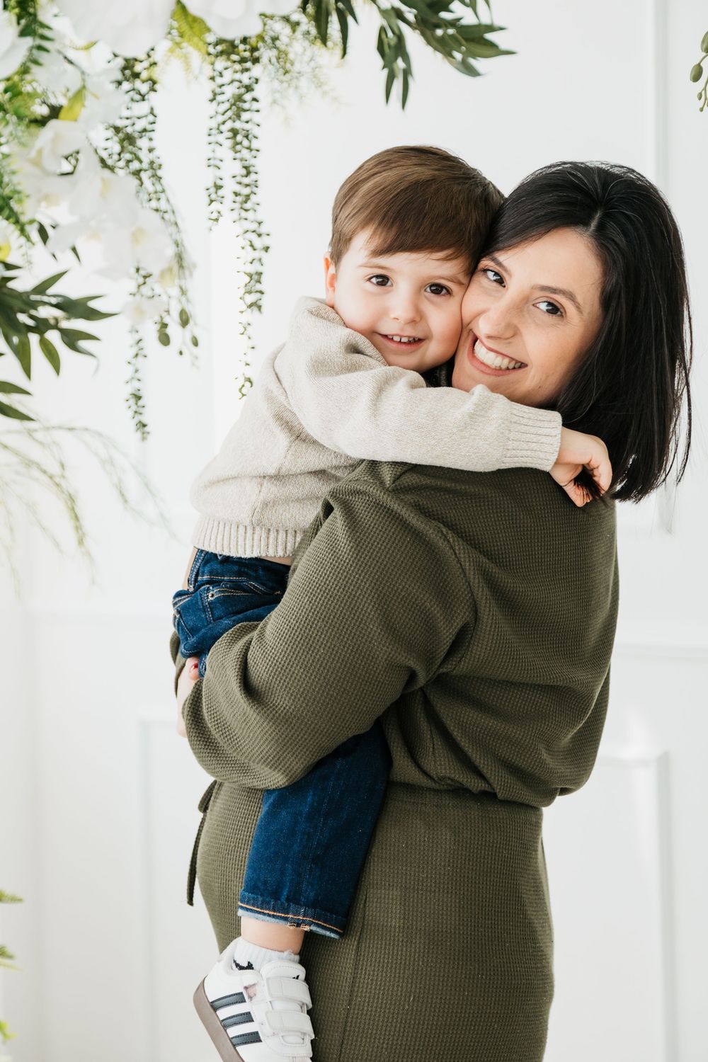 A tender embrace between parent and child against a white backdrop with delicate green foliage decor.