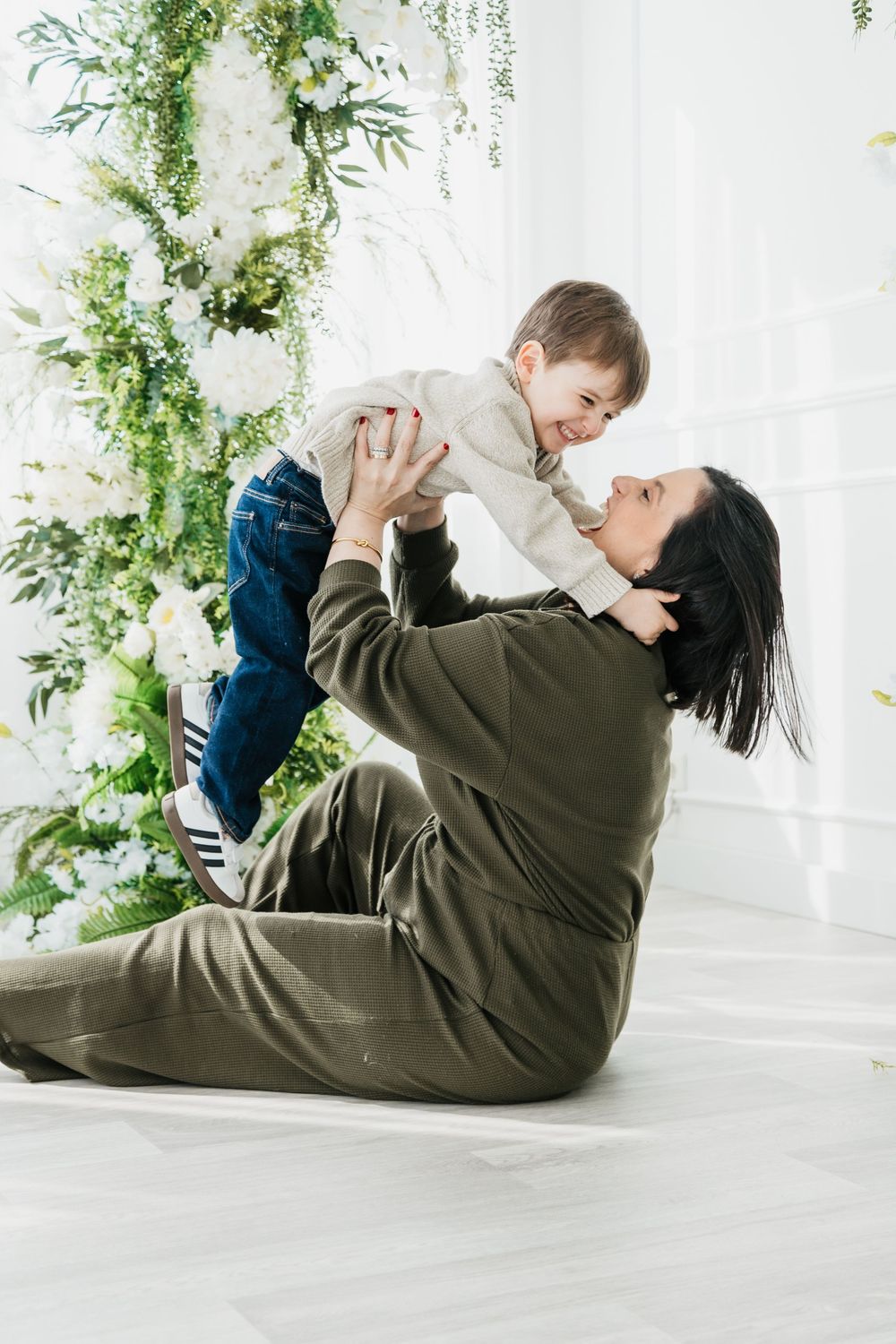 A tender family moment captured in a bright white studio with hanging greenery and floral decorations.