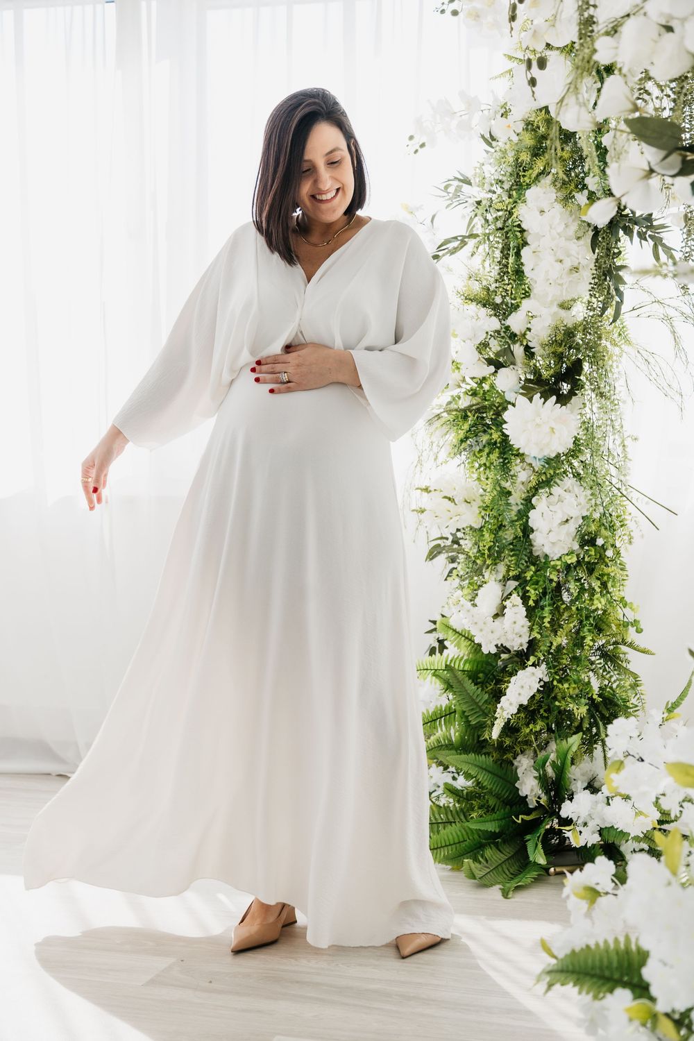 A person in a flowing white dress poses gracefully against a backdrop of white flowers and green foliage.