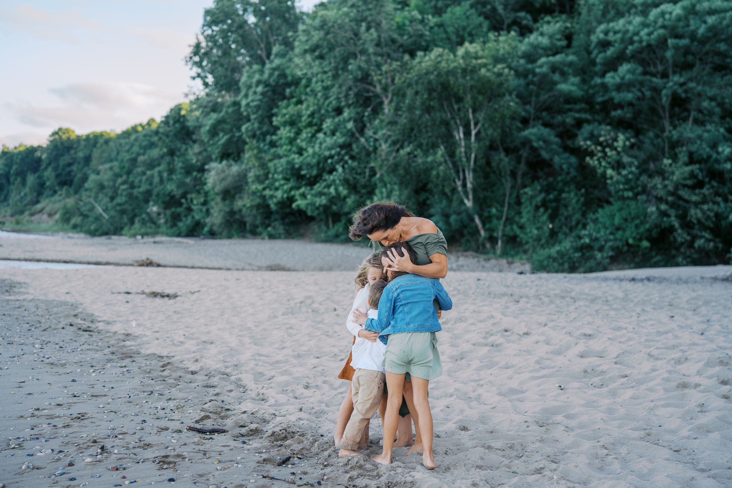 Three people embrace while standing on a sandy beach near a tree-lined shore at dusk.