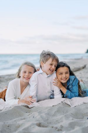 Three children lying together on a sandy beach, laughing and smiling while wearing white and blue clothing.