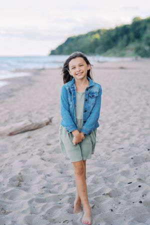 Young girl in denim jacket smiles while standing barefoot on a sandy beach near ocean cliffs.