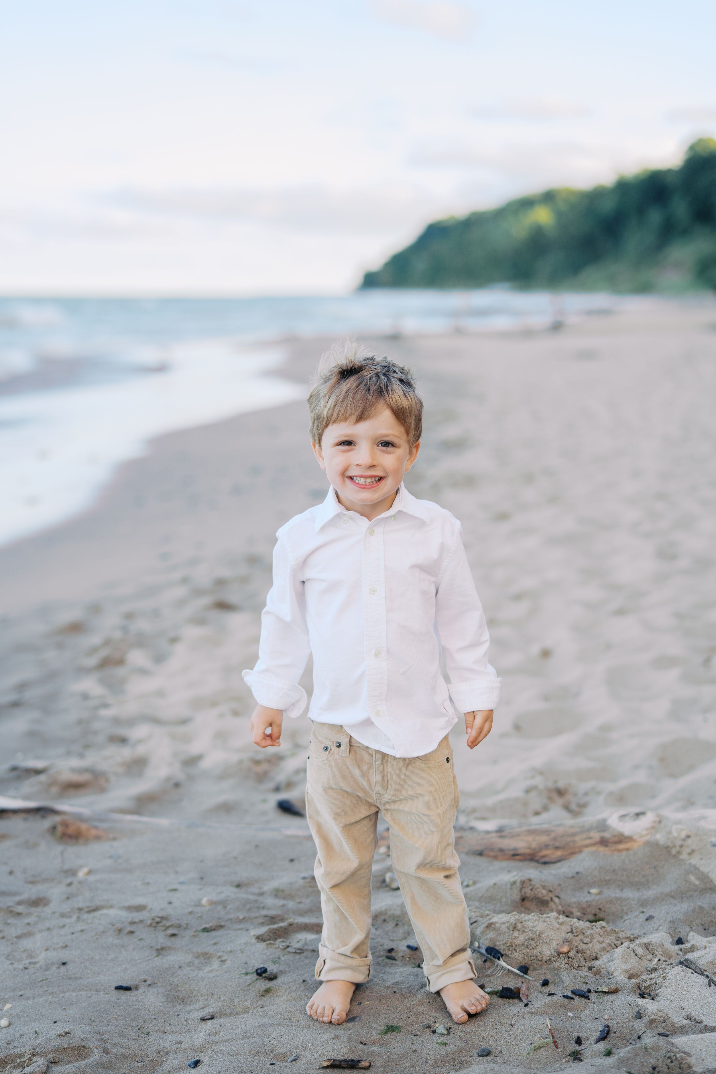 A young child in a white shirt and khaki pants plays barefoot on a sandy beach with bluffs in the background.