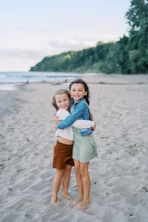Children share warm embraces while playing barefoot on a sandy beach near trees and water.