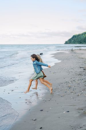A joyful figure in denim runs barefoot along a misty beach shoreline at dusk with forested cliffs in the distance.