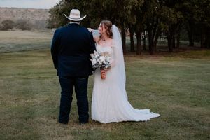 Bride in white gown and veil holds bouquet while groom in navy suit and cowboy hat touches her shoulder outdoors.
