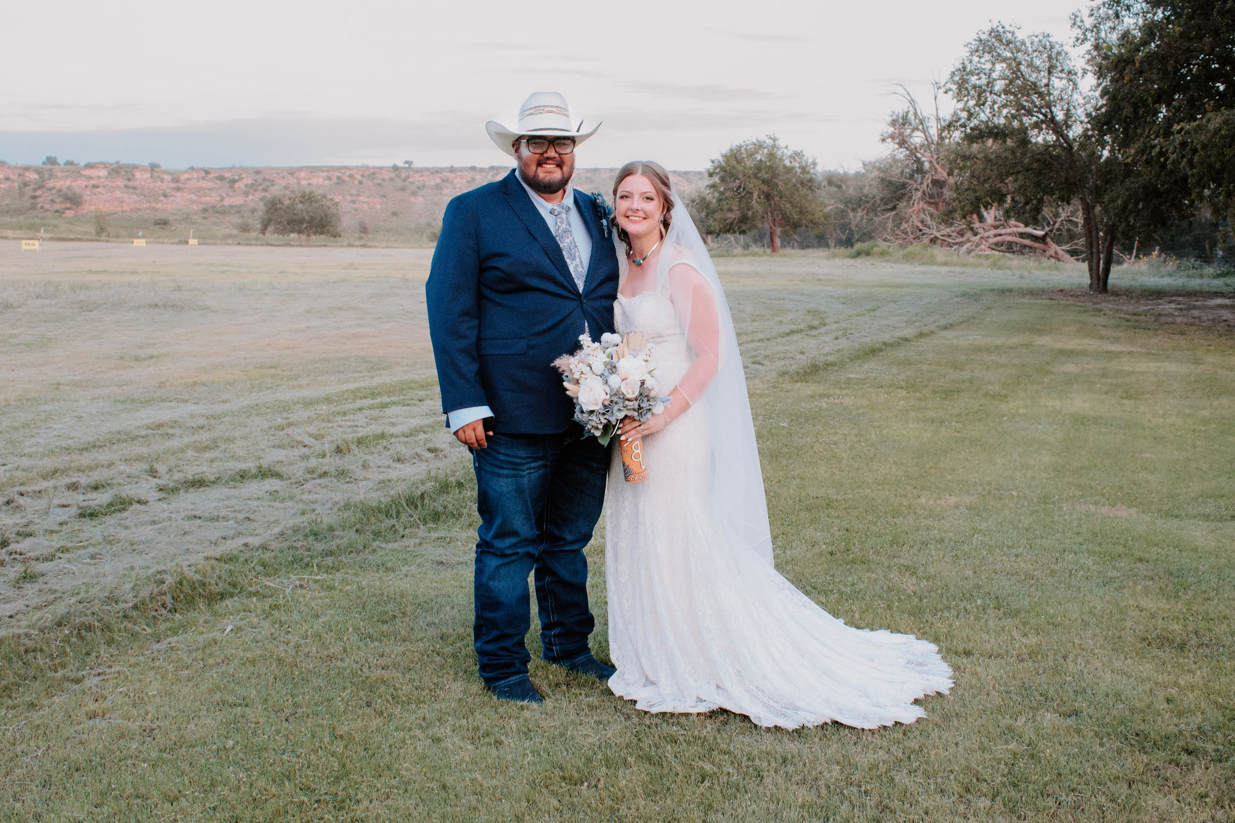 Bride and groom posing outdoors on green lawn, groom in blue suit and cowboy hat, bride in white gown holding bouquet.