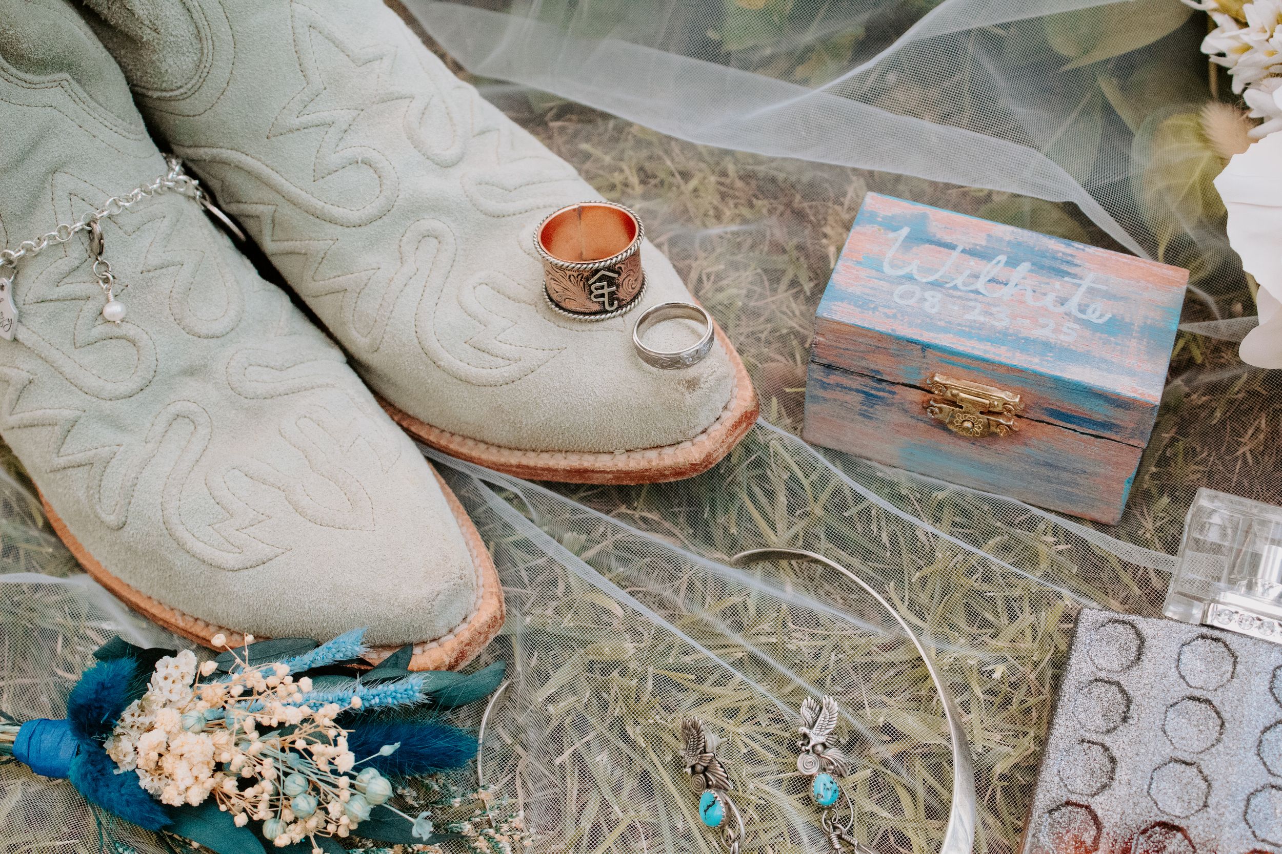 Rustic wedding flat lay with cowboy boots, gold rings, a decorative box, blue bouquet, and veil on hay.