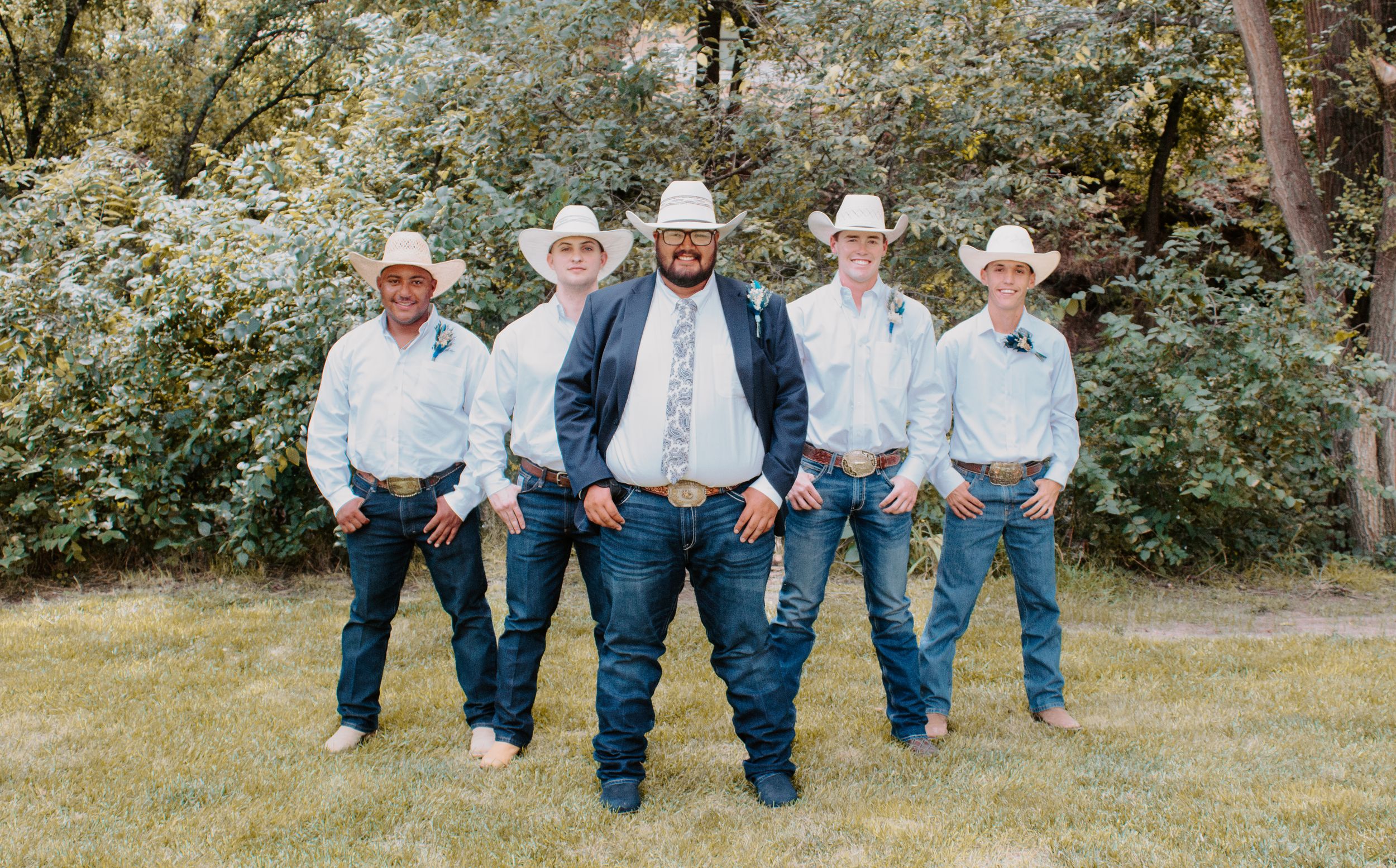Five men in white cowboy hats and jeans pose outdoors on grass for a western-themed wedding party photo.
