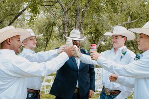 Groomsmen in white shirts and cowboy hats toasting drinks outdoors at a rustic wedding celebration.