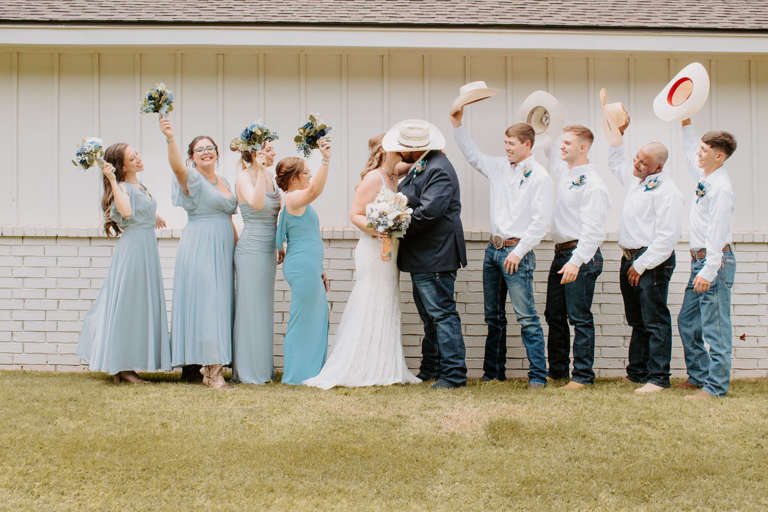 Wedding party celebrates as bride and groom kiss, bridesmaids in blue dresses, groomsmen in cowboy hats raising them in cheer.