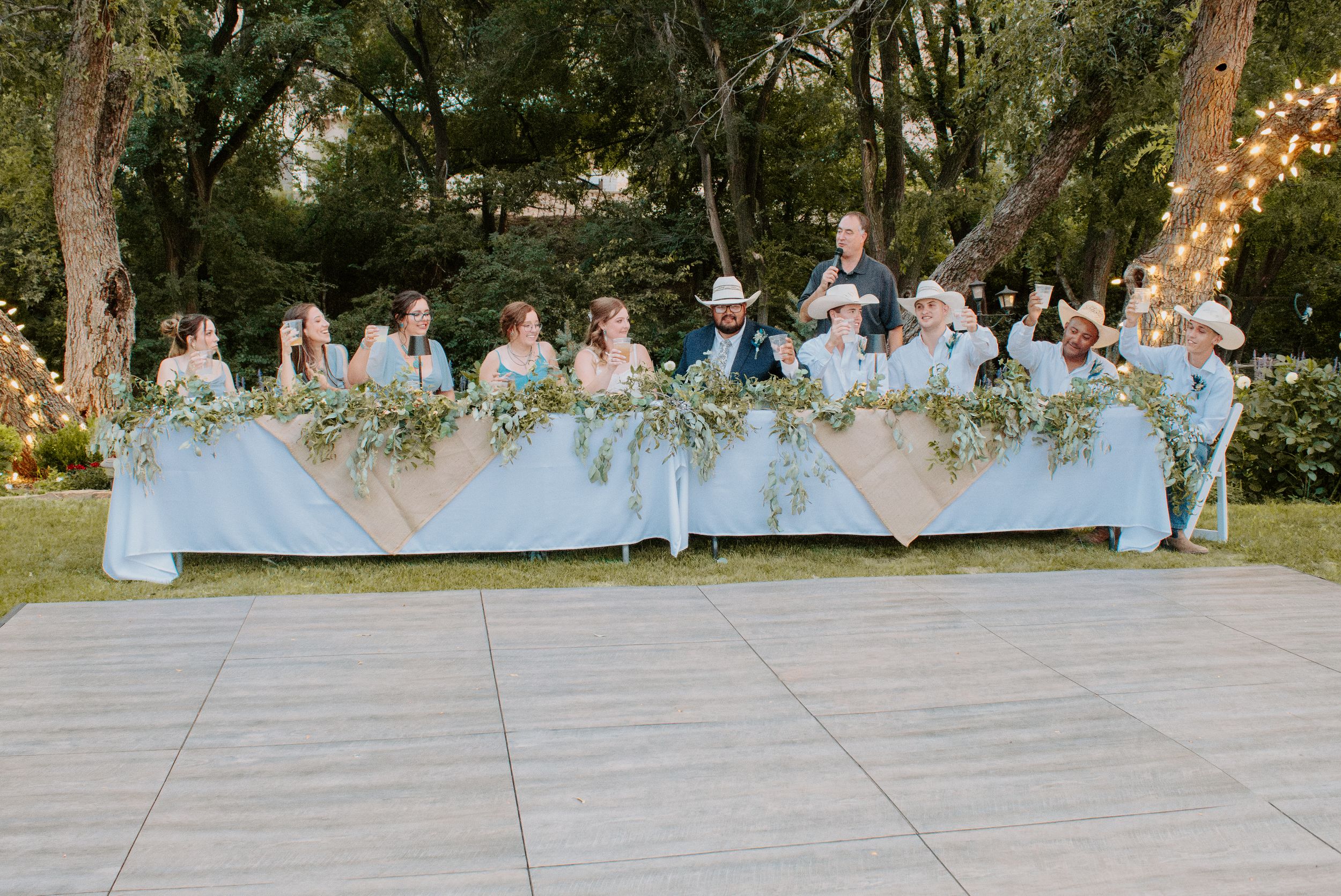 Outdoor wedding party seated at a long decorated table with greenery garlands, string lights, and blue linens in a garden setting.