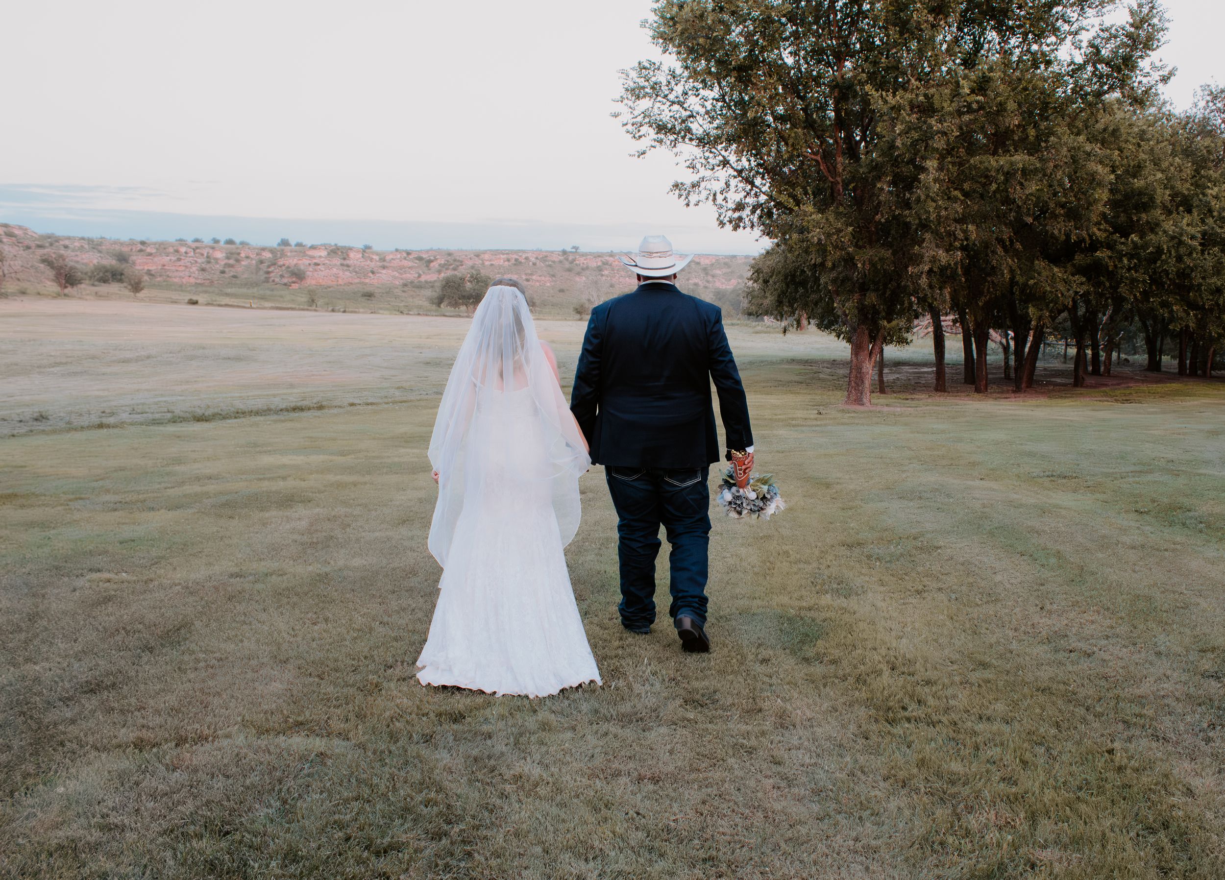 A bride in a white gown and veil walks with a man in a suit across a green lawn at an outdoor wedding venue.