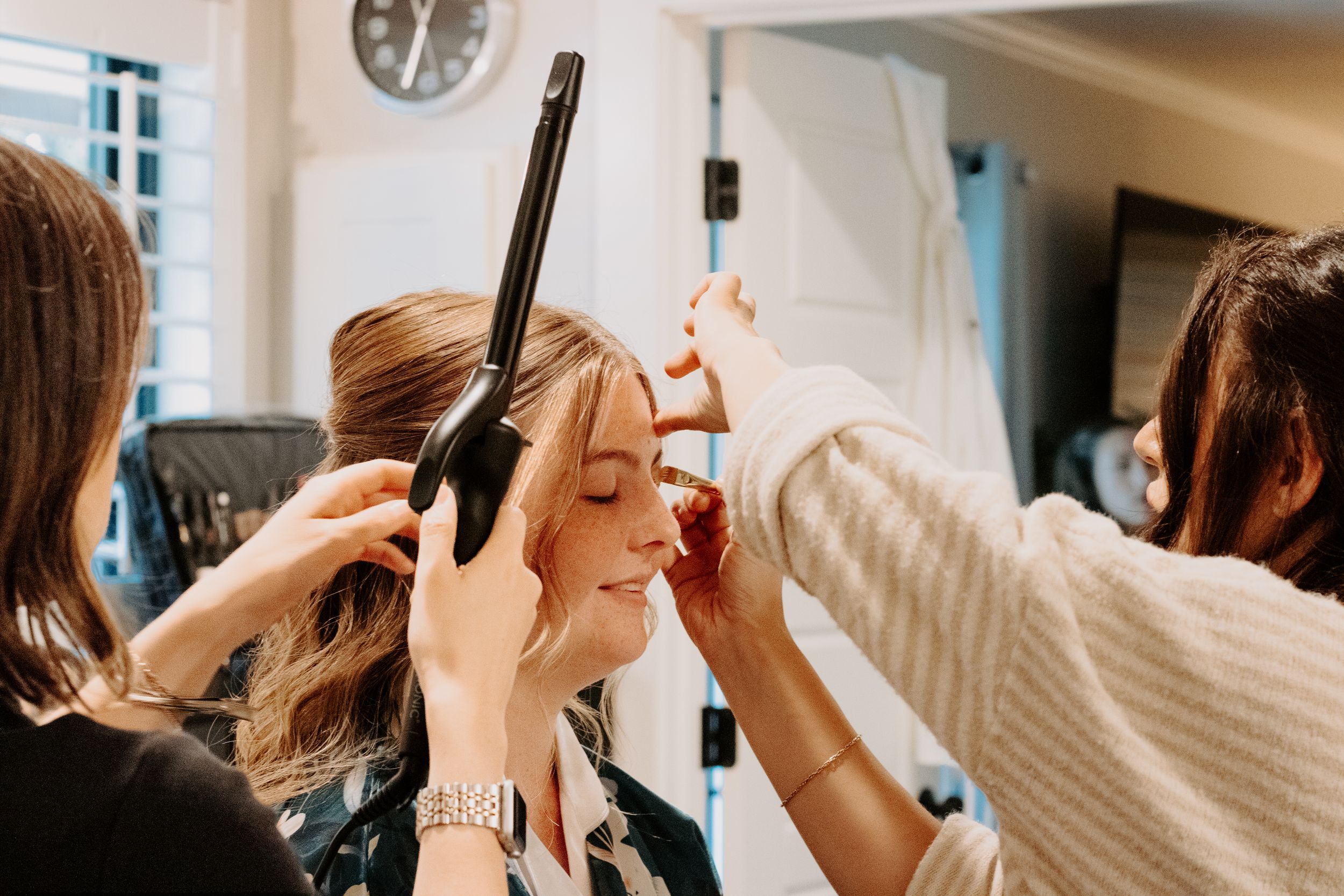 Two stylists curl and apply makeup to a smiling blonde woman during a bridal beauty preparation session indoors.