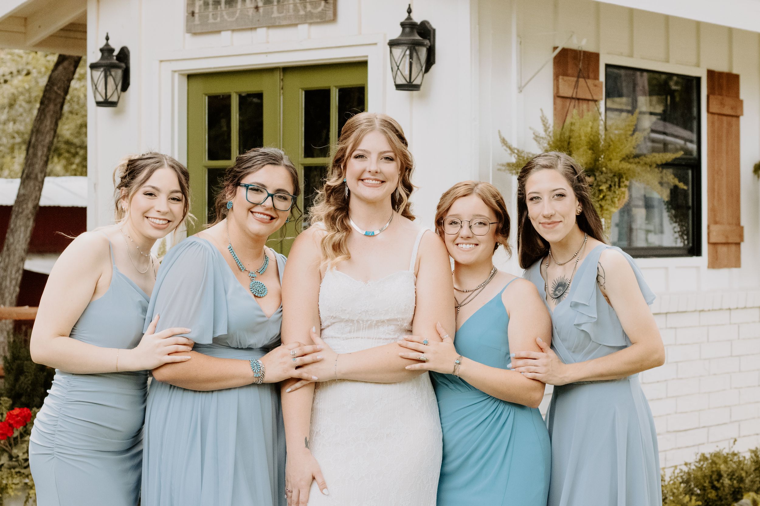 Bride in white gown poses with four bridesmaids in dusty blue dresses outside a white building.