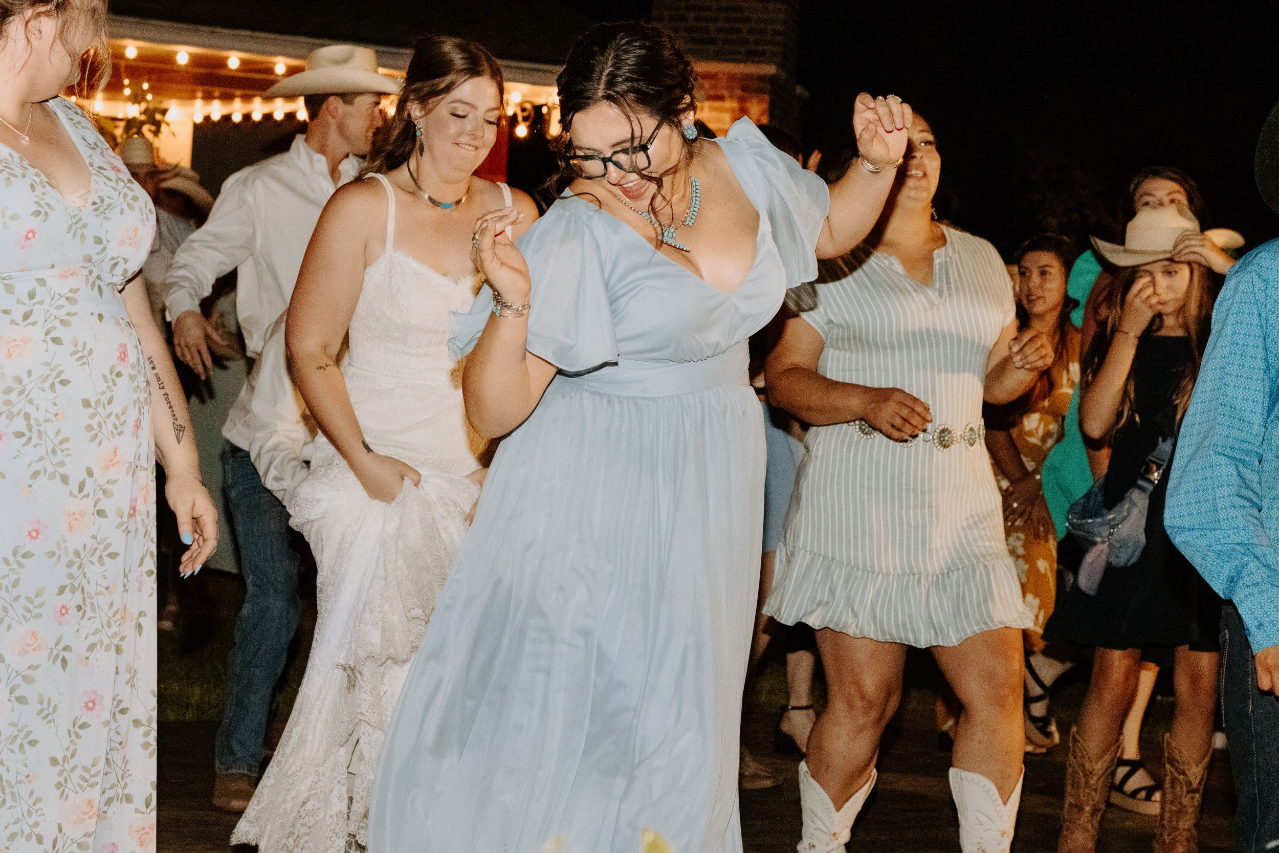 Women in elegant dresses dance joyfully at an outdoor evening wedding reception with string lights.