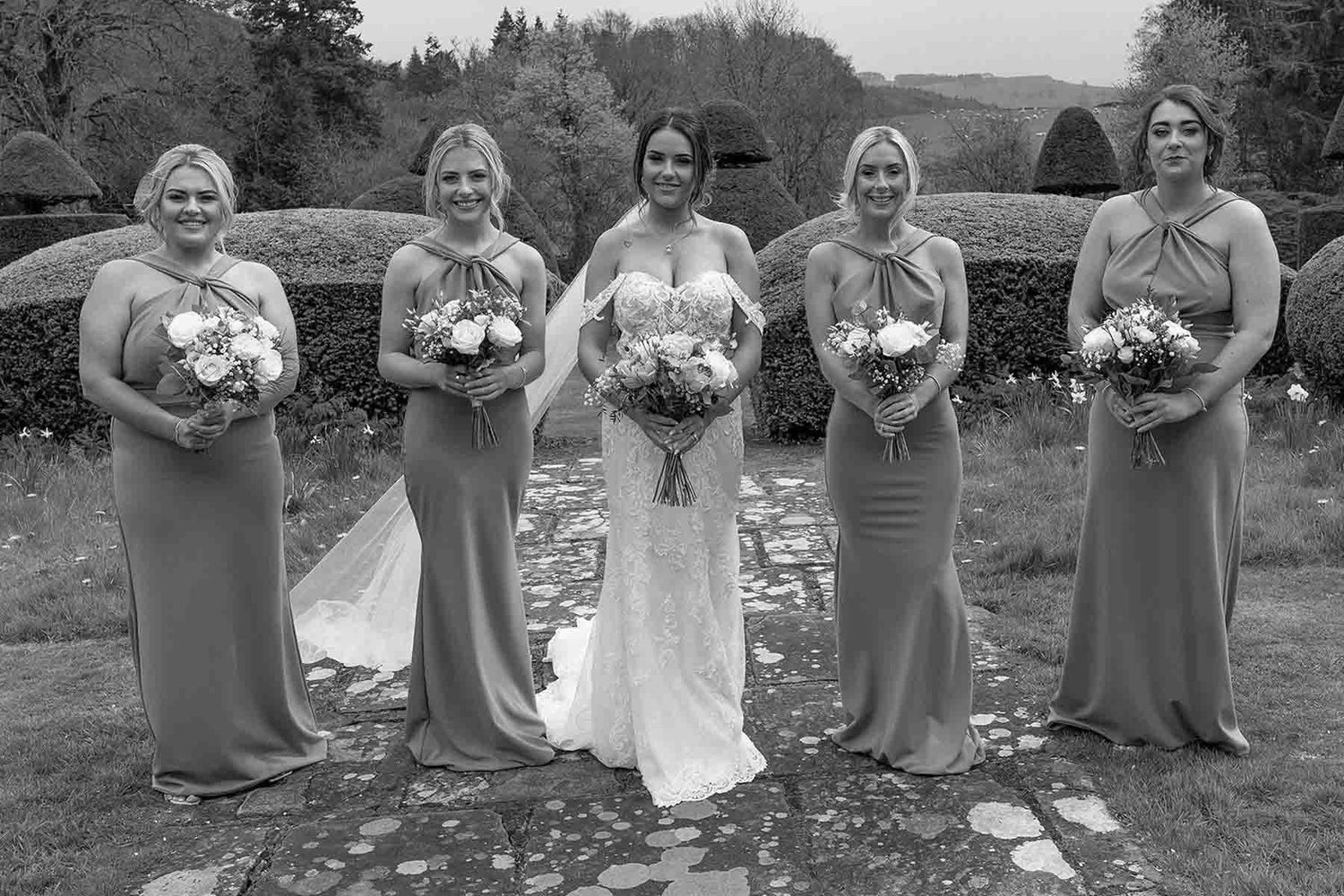 Black and white wedding party photo showing a bride and four bridesmaids in elegant gowns holding bouquets outdoors.