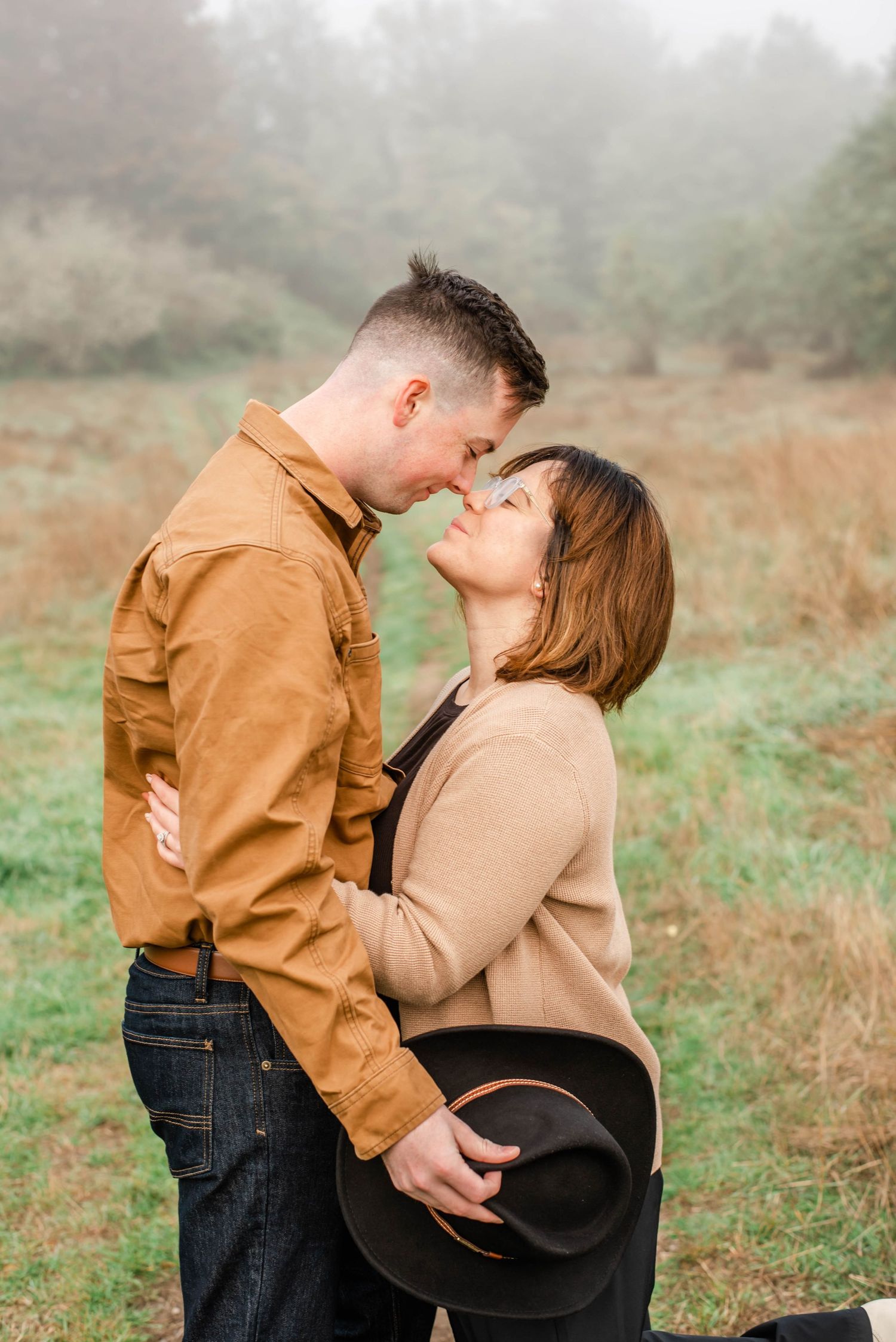 A couple shares a tender moment outdoors in a field during golden hour, wearing casual attire.