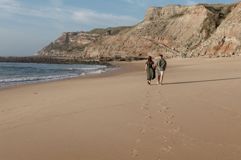 Two people walking along a sandy beach with cliffs in the background.