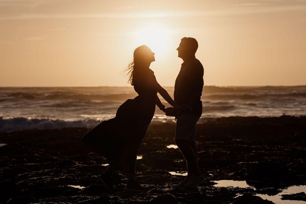 Two silhouetted figures stand together on a beach at sunset, the ocean waves crashing in the background.