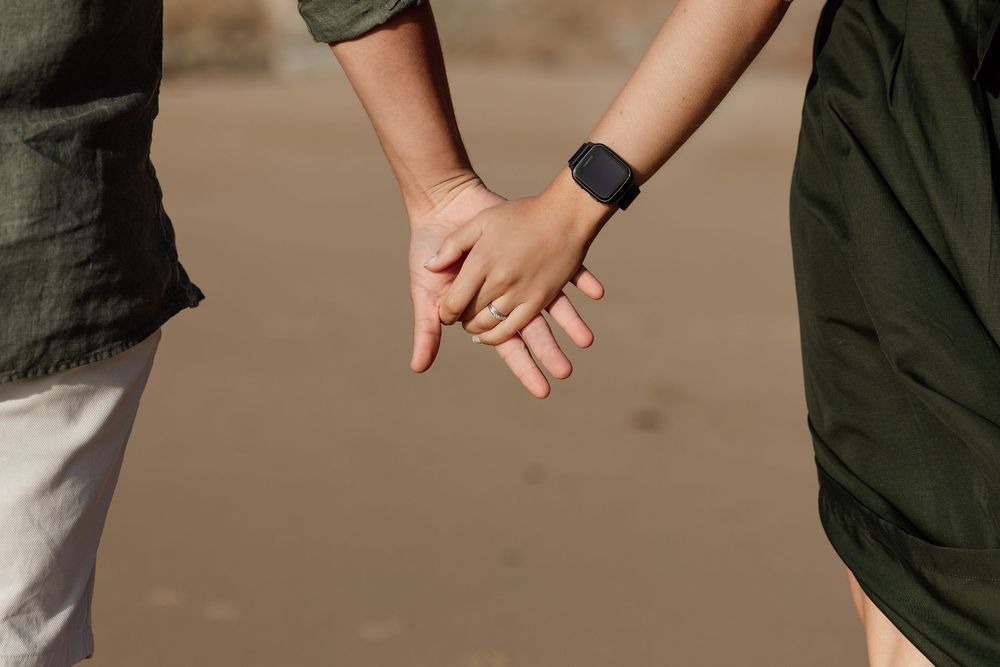 Two hands holding each other in a close embrace on a sandy beach.