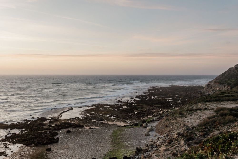 A scenic coastal landscape with rocky shoreline, crashing waves, and a calm ocean horizon under a cloudy sky.