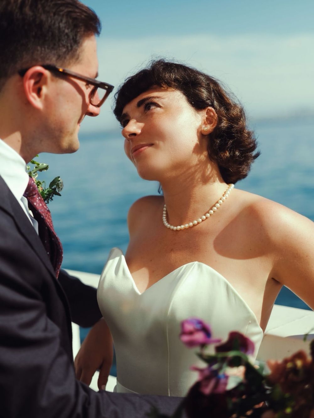 A bride in a strapless white gown and pearl necklace gazes at her groom in a suit near the ocean.