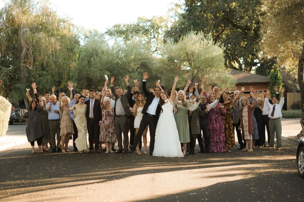 Joyful wedding group photo with bride and groom surrounded by cheering guests outdoors among trees.