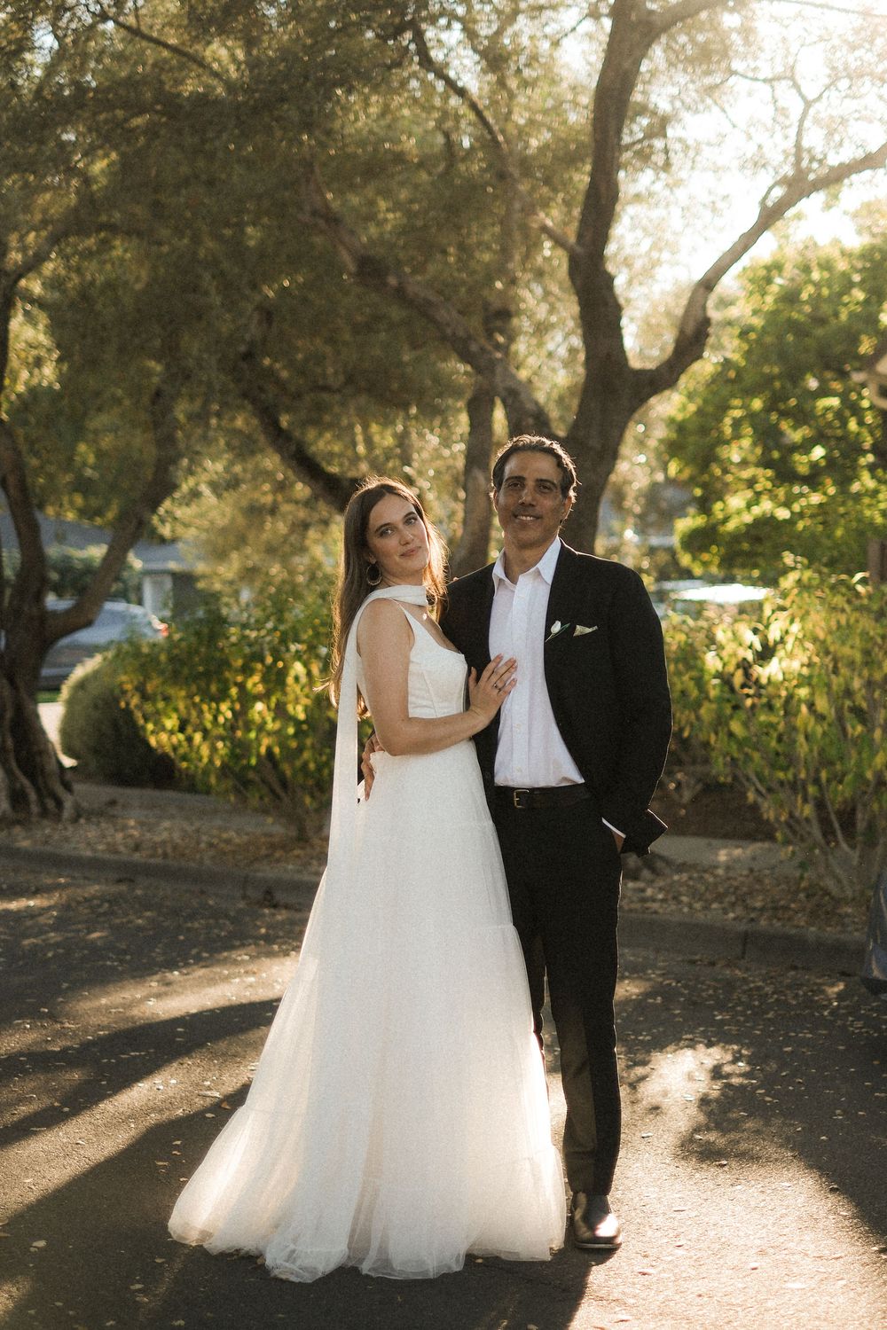Bride in white gown and groom in black suit pose together outdoors among trees in golden sunlight.