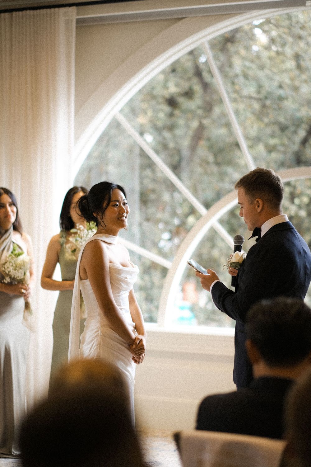 Bride and groom exchange vows indoors by arched window, bridesmaids in white holding bouquets during elegant wedding ceremony.