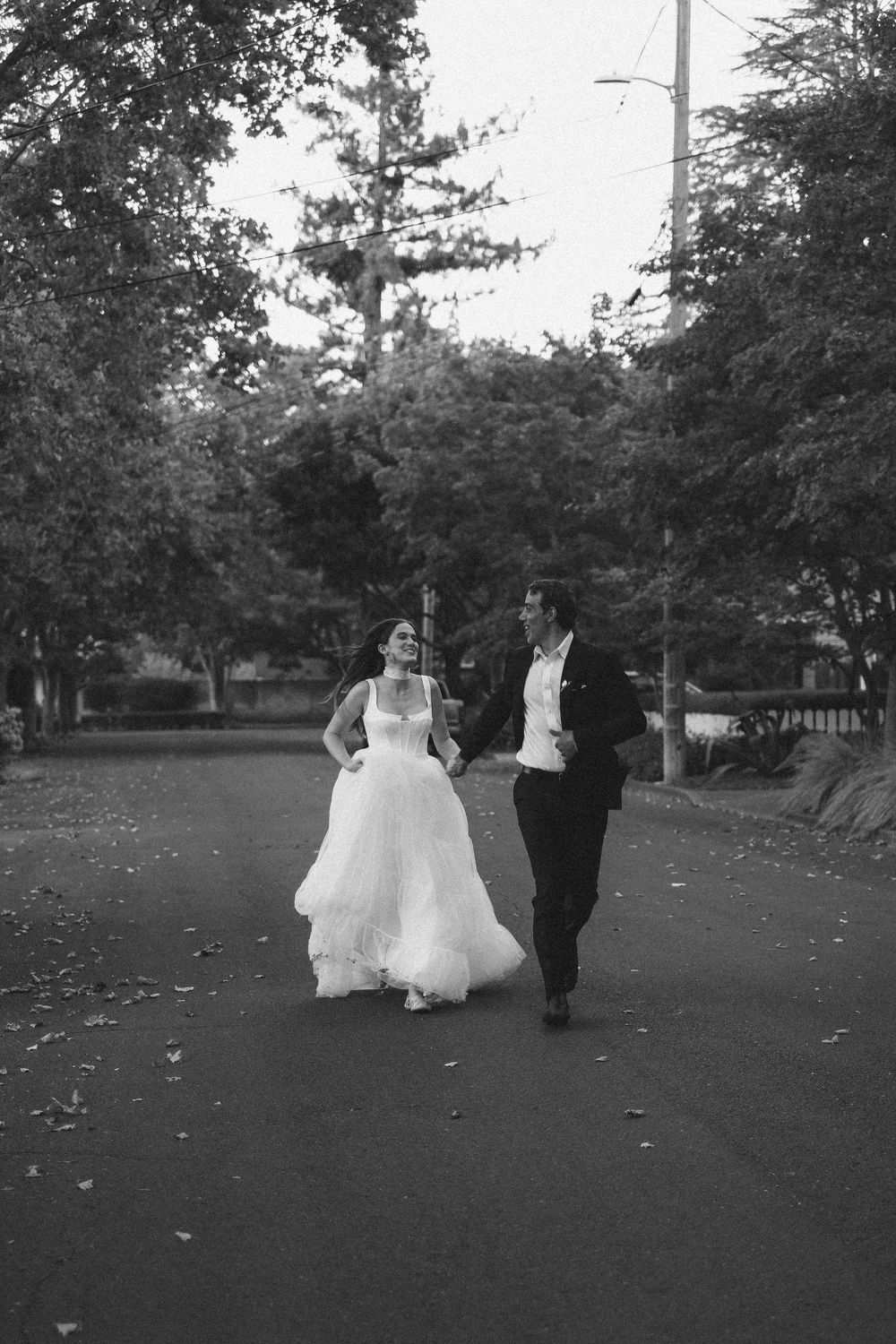 Black and white photo of a bride and groom walking hand in hand down a tree-lined path on their wedding day.