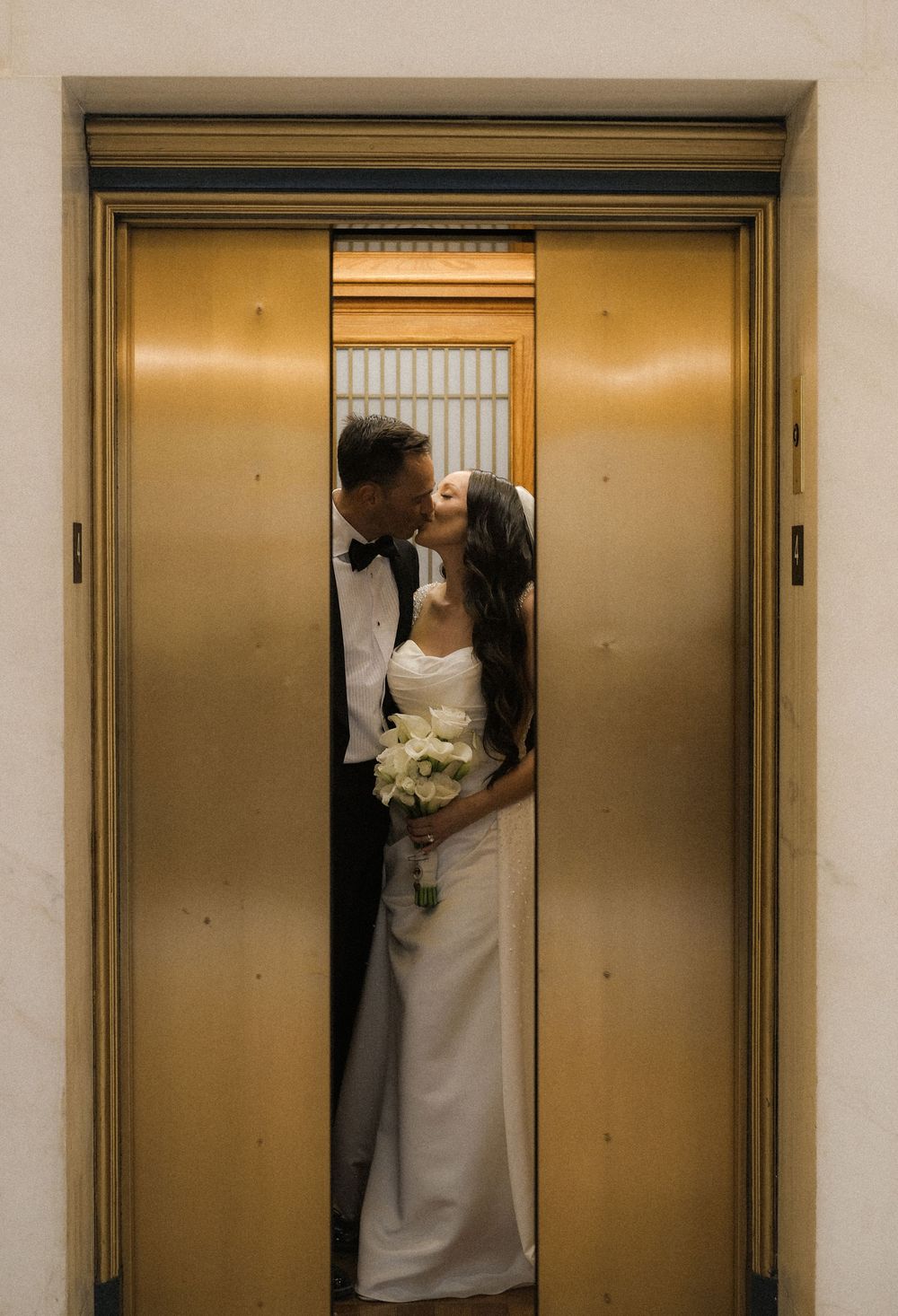 Bride and groom sharing a romantic kiss inside a golden elevator, bride holding white bouquet in elegant wedding attire.