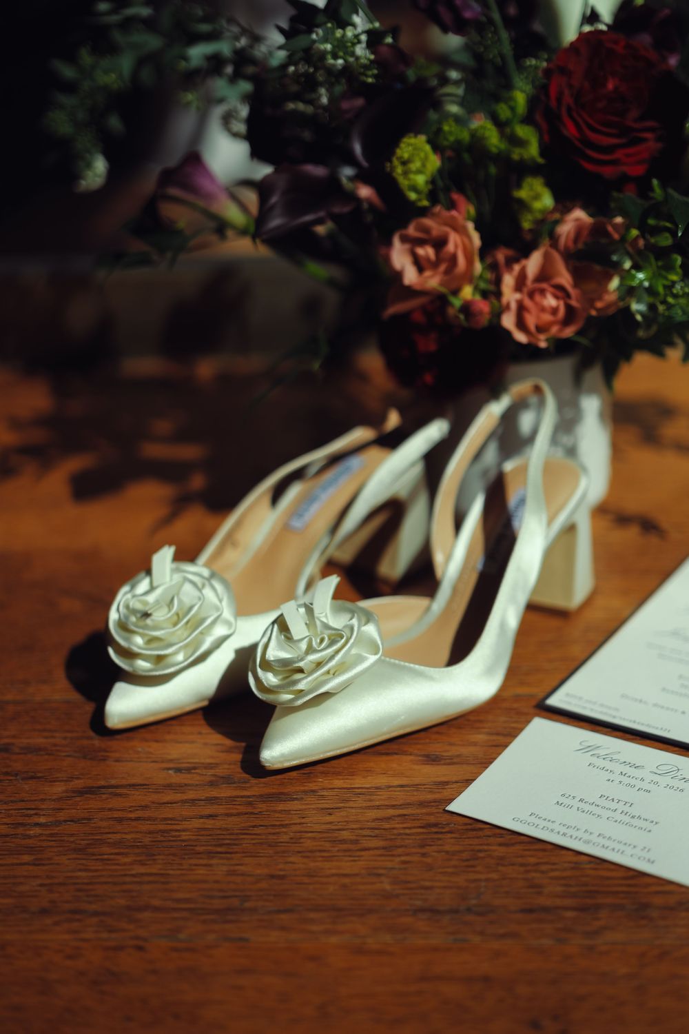 White satin bridal heels with rose detail on wooden table beside wedding bouquet and invitation cards.
