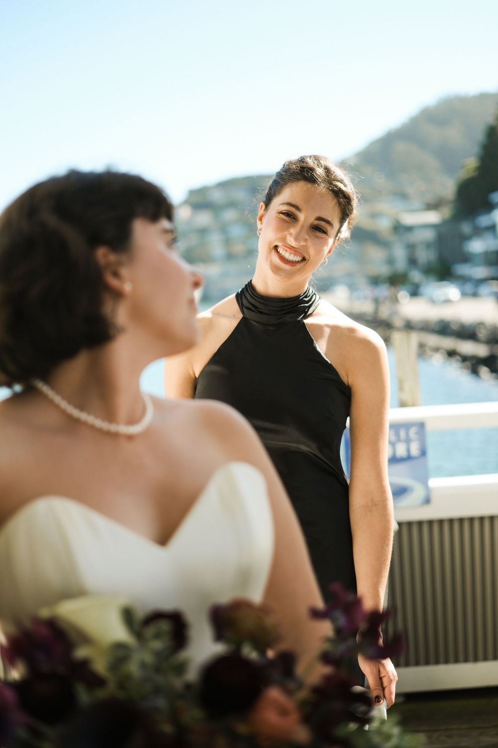 Smiling bridesmaid in black halter dress stands beside bride in white gown at waterfront wedding venue.