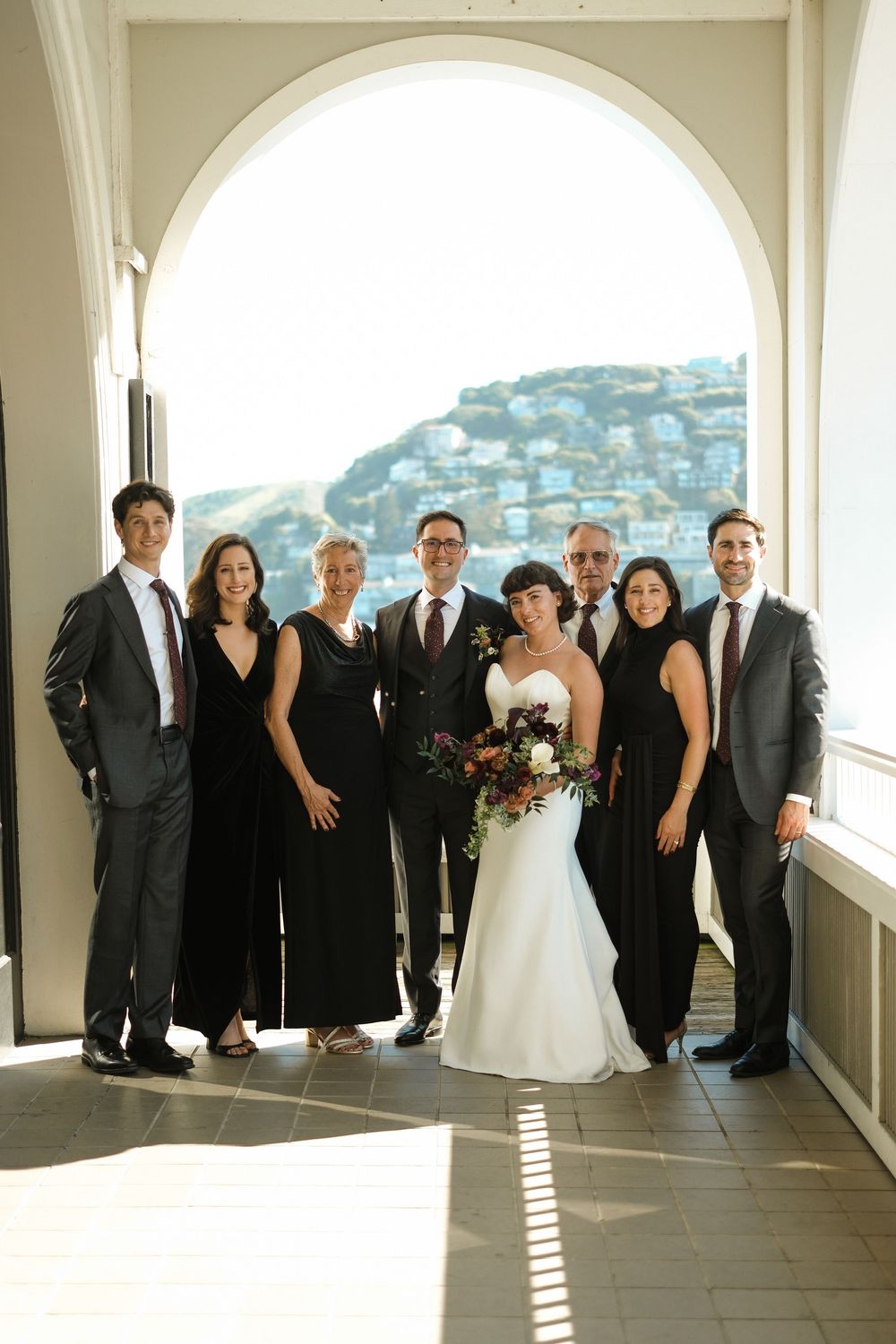 Wedding party posing under arched window with scenic hillside view, bride in white gown holding bouquet surrounded by guests in black.