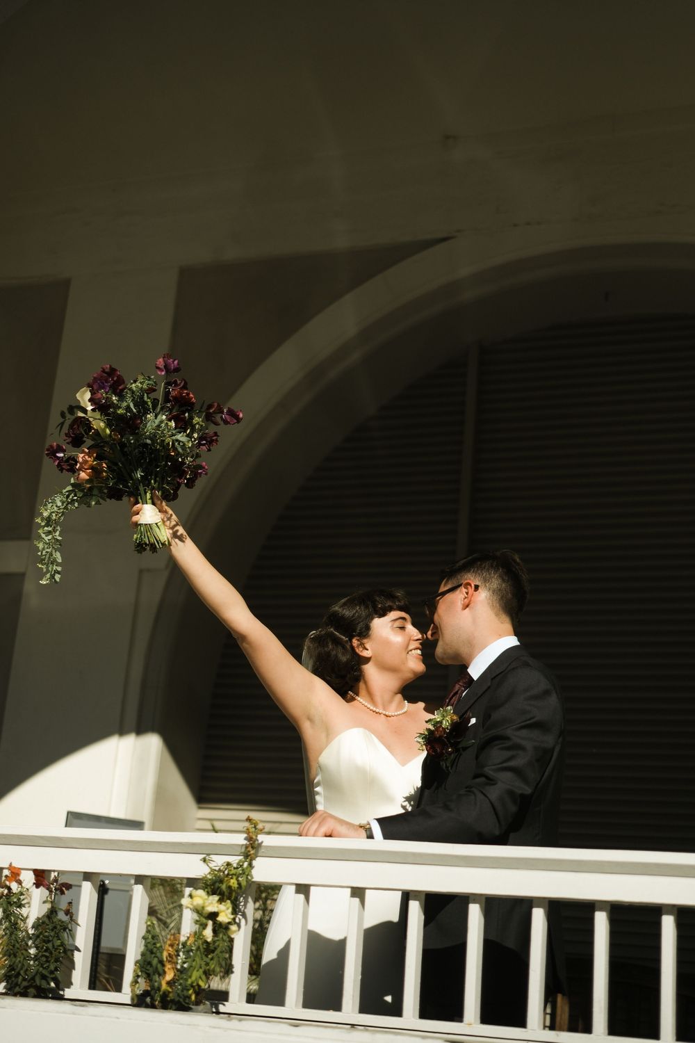 Joyful bride raises bouquet on balcony while groom kisses her cheek in wedding attire.
