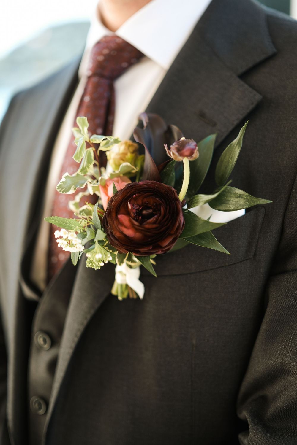 Elegant groom wearing dark suit with burgundy tie and floral boutonniere featuring deep red ranunculus and greenery.